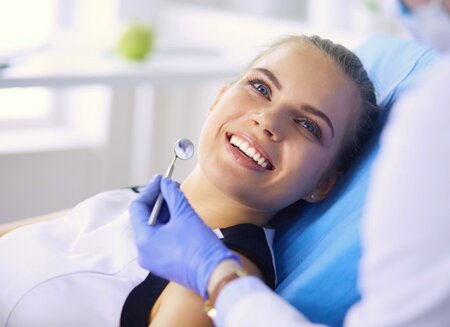 blond girl smiling sitting in dental exam chair, dental mirror near mouth being held by hygienist, teeth cleaning Boca Raton, FL dentist